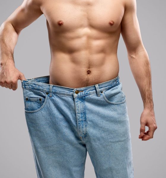 Anonymous shirtless male with muscular torso pulling huge jeans after successful weight loss against gray background