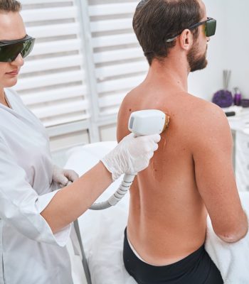 Young dark-haired female doctor in safety goggles and disposable nitrile gloves performing a cosmetic procedure with a laser device