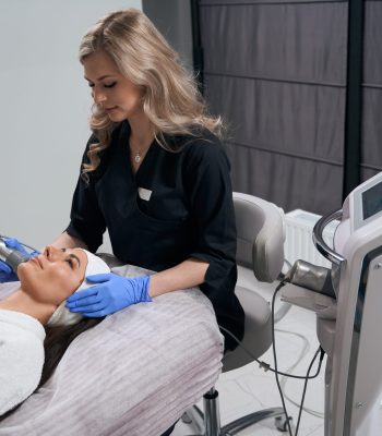 Beautiful female lying on couch in salon, cosmetologist in protective gloves doing RF lifting for face