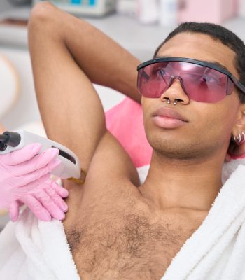 Male in safety goggles lying supine during armpit hair removal conducted by cosmetologist in nitrile gloves